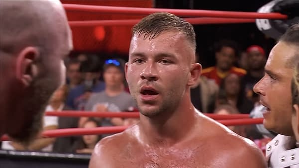 Daniel Garcia stands in a wrestling ring, visibly torn between two individuals, with an audience in the background. His expression conveys conflict and uncertainty.