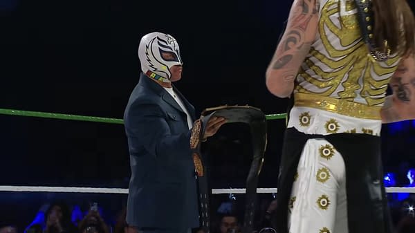 Rey Mysterio, wearing a signature mask, presents Dominik Mysterio with the AAA Mega Championship belt in a wrestling ring at WWE x AAA Worlds Collide. The scene captures a ceremonial moment between the two wrestlers.