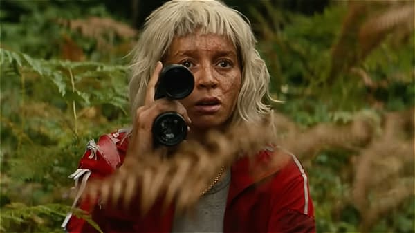 Erin Kellyman as Jimmy Ink in In 28 Years Later: The Bone Temple. A young woman with short, light-colored hair and freckles gazes intently through binoculars, surrounded by greenery and ferns. She wears a red jacket and appears focused on something in the distance.