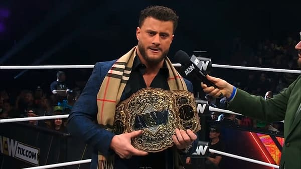 A wrestler holds the AEW World Championship belt while being interviewed by another individual, dressed in a blazer and a plaid scarf. The wrestling ring is visible in the background with an audience.