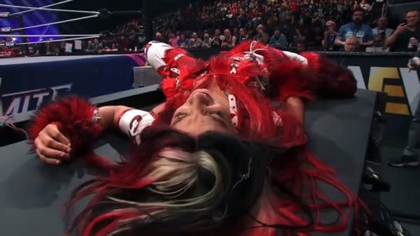A wrestler lies on a table, looking defeated, with vibrant red hair and a flashy outfit during AEW Dynamite's New Year's Smash event. The audience can be seen in the background, reacting to the scene.