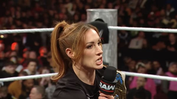 Becky Lynch stands in a wrestling ring on WWE Raw, holding a championship belt while speaking into a microphone. The audience is visible in the background, showcasing their enthusiastic reactions.