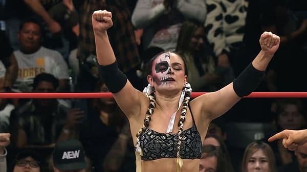 Thunder Rosa, an AEW wrestler, stands triumphantly with her arms raised, showcasing her striking face paint and braided hair, while fans cheer in the background.