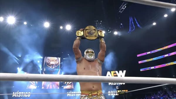 Místico, wearing a mask and wrestling attire, raises his newly won Trios Championship above his head, celebrating in the spotlight while a cheering crowd surrounds him.
