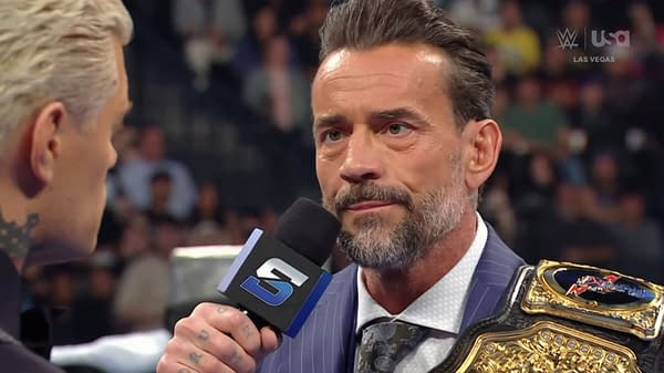 CM Punk speaks directly to Cody Rhodes while holding a championship belt, with a crowd visible in the background. Both wrestlers are engaged in a tense moment during WWE SmackDown.