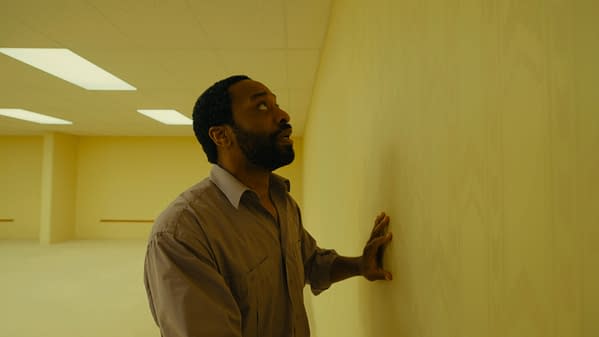 A man stands in a yellow-lit, empty Backrooms-style hallway, touching the wall and looking upward with a worried expression.
