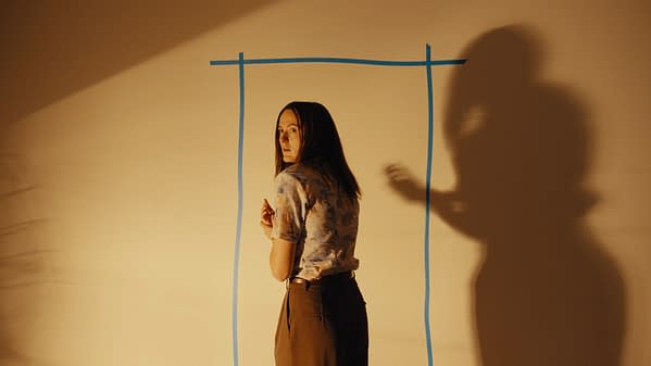 A woman stands in a taped-off doorway-like rectangle on a blank wall, glancing back nervously as a large shadow looms beside her in a eerie Backrooms-style scene.