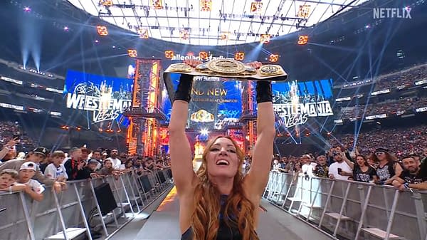 A female wrestler holds up two championship belts triumphantly in a wrestling ring arena, surrounded by an excited crowd. The background features a large display screen announcing the WrestleMania event.