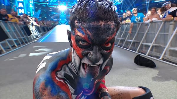 A wrestler with face paint in a striking design featuring black, red, and blue colors, showing intense facial expression and sticking out his tongue. The backdrop includes a cheering crowd and bright stage lights.