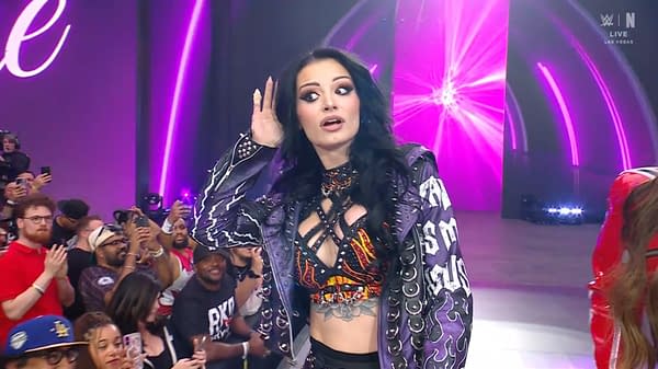 A wrestler with long black hair and striking makeup wears a studded purple jacket over a colorful top, reacting to the crowd in a lively arena filled with fans.