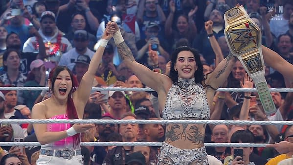 Two women celebrate in a wrestling ring at WrestleMania, with one holding a championship belt and both smiling and raising their arms in victory. The crowd is enthusiastic in the background.