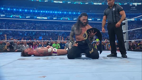 A wrestlers kneel on the mat, holding a championship belt, while another lies defeated nearby. The audience in the background is cheering and cheering, capturing the energy of a major wrestling event.
