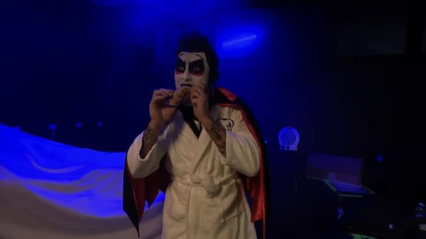 Danhausen, in face paint and a robe, eats a snack under blue lighting on WWE SmackDown.