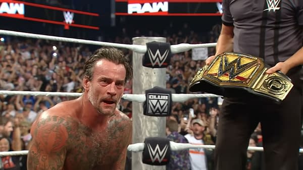 CM Punk kneels in the WWE ring while a referee hands over the WWE Championship after his Raw victory. The crowd blurs in the background as the title is presented at ringside.