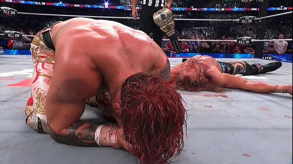 Will Ospreay and Kenny Omega lie exhausted on the mat after the AEW All In London main event, with a championship belt visible in the ring. The crowd watches in the background.