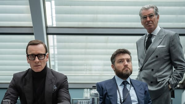 Three men are seated in a modern conference room; one man in the foreground wears glasses and a turtleneck, the second man in a suit appears contemplative, and the third man in the background stands with a serious expression. Black Bag Review: Another Slick And Stylish Thriller From Soderbergh.