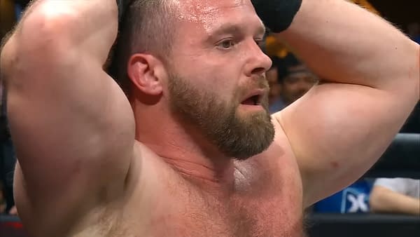 A close-up of a muscular man, Cash Wheeler of FTR, with a beard, showing an intense expression as he holds his head in a wrestling ring, during a moment of tension.