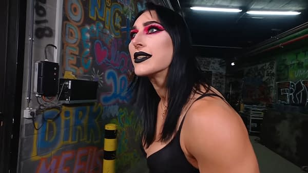 Rhea Ripley, a professional wrestler, is shown backstage at WWE Raw in Brussels, wearing striking makeup with bold pink and black colors. The background features colorful graffiti art, adding to the vibrant setting.