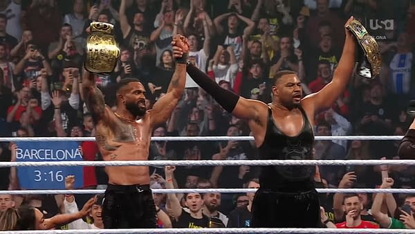 Two wrestlers from the Street Profits celebrate in the ring after winning a championship, with fans cheering in the background. One holds a championship belt aloft while the other raises his arm triumphantly.