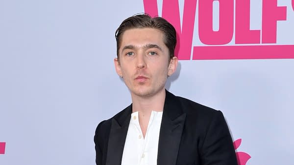 A man with short brown hair and earrings poses confidently in front of a light background featuring bright pink text, indicating a promotional event or premiere.