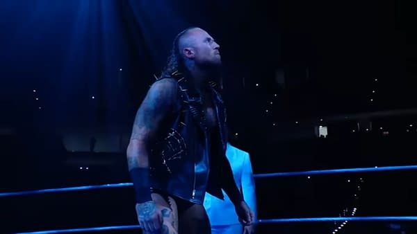 A wrestler, Aleister Black, with a spiked black vest and tattoos stands in a wrestling ring under dramatic blue lighting. He gazes upwards, appearing intense and ready to make a statement.