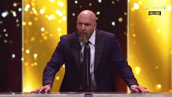 Triple H stands at a podium, wearing a dark suit and tie, as he accepts his induction into the 2025 WWE Hall of Fame. A bright, golden backdrop enhances the celebratory atmosphere.