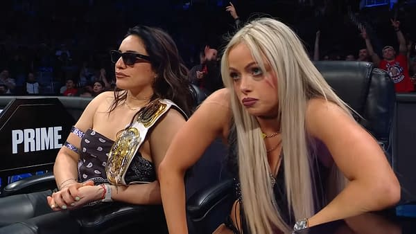 WWE Women's Tag Champions Raquel Rodriguez and Liv Morgan sit attentively during a tag team gauntlet match on WWE SmackDown. Raquel wears sunglasses and a championship belt, while Liv appears focused and engaged.