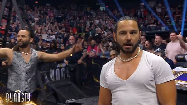 Two members of the Young Bucks appear onstage during an AEW event, with a cheering crowd behind them, as one gestures dramatically. The atmosphere is charged with excitement.