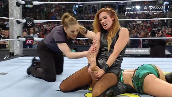 Becky Lynch applies pressure on Lyra Valkyrie's arm while an official intervenes during a WWE match. The background features a cheering crowd and wrestling ring elements.