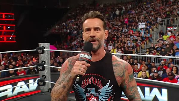 A wrestler with a tattooed arm, CM Punk, speaks into a microphone while standing in a wrestling ring on WWE Raw. The audience is visible in the background, showing excitement during the live event.