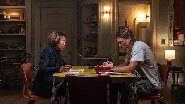 A tense conversation between two characters at a yellow table, featuring a woman in a navy blazer and a man in a casual t-shirt, both focused on papers in front of them. The background shows bookshelves and a cozy apartment setting.