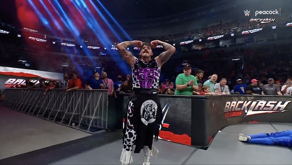 Dominik Mysterio flexes his muscles confidently after executing a successful maneuver during a WWE Backlash event, surrounded by cheering fans and colorful stage lights.