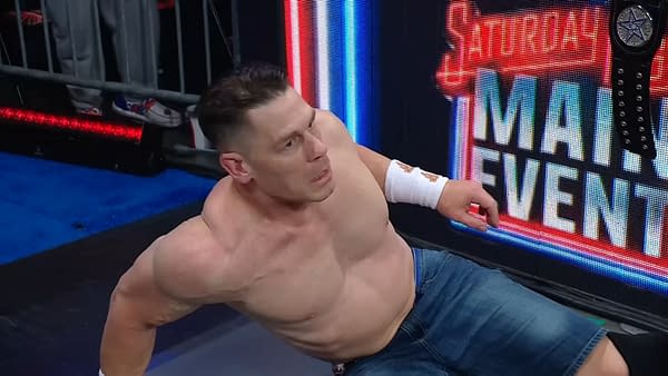John Cena, with a shocked expression, is seated on the floor in front of a backdrop featuring the WWE Saturday Night's Main Event logo. He is shirtless and wearing denim shorts, with visible muscles and a bandage on his arm.