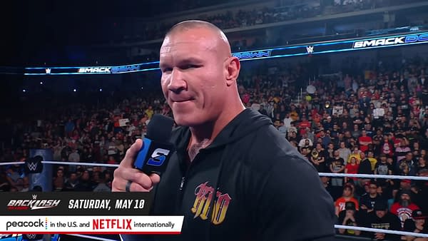 Randy Orton delivers a promo on WWE SmackDown, speaking into a microphone in front of a cheering crowd. He wears a black hoodie with a colorful design, emphasizing an intense moment in the ring.