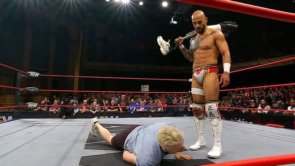 A muscular wrestler, Ricochet, stands triumphantly over Zach Gowen, who is on the mat. Ricochet holds Gowen's own prosthetic leg as a symbolic act during an AEW Collision event.
