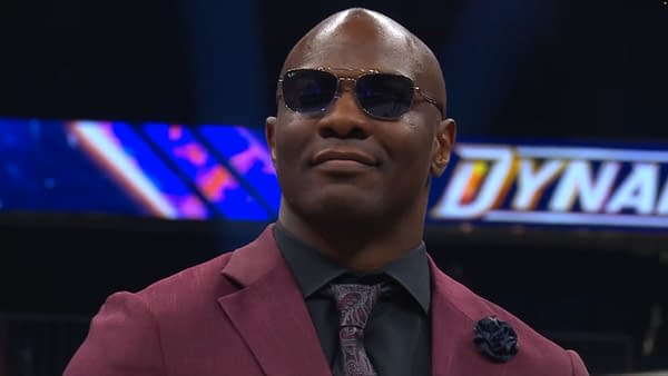 A confident Shelton Benjamin wearing sunglasses and a stylish dark suit with a patterned tie, standing on the set of AEW Dynamite. The background features the AEW Dynamite logo in vibrant colors.