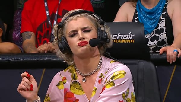 A commentator with blonde hair and bold makeup, dressed in a floral blouse, looks skeptically while speaking into a microphone, headset on. The background shows a diverse audience at an AEW event, highlighting a mix of expressions from fans.
