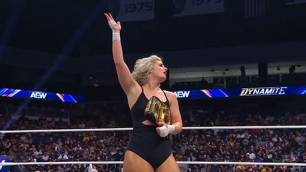 Toni Storm, wearing a black wrestling outfit, celebrates her victory in the ring, holding a championship belt above her head with her right hand raised. The crowd in the background is cheering her on.