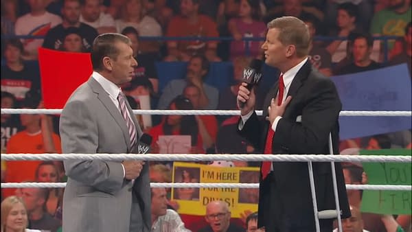Vince McMahon, in a gray suit, stands on a wrestling ring with John Laurinaitis, who is wearing a black suit and using crutches. Fans holding colorful signs are visible in the background.