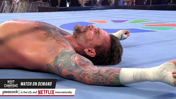 A defeated wrestler lies on the wrestling ring floor, showcasing tattoos on his arms and a weary expression. It is CM Punk in the aftermath of his match with John Cena at WWE Night of Champions in Saudi Arabia.