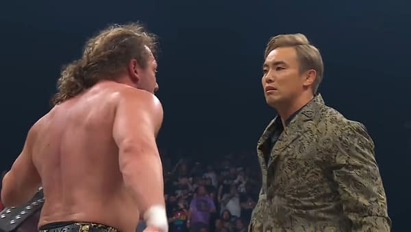 Kenny Omega and Kazuchika Okada face each other intensely during AEW Fyter Fest, capturing a moment of confrontation between two wrestling stars. Omega, with his long hair and muscular physique, faces Okada, who is dressed in an ornate jacket, emphasizing the tension of their rivalry.
