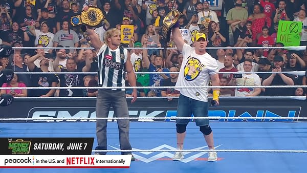 Logan Paul and John Cena stand in the WWE ring, both holding championship belts aloft, surrounded by an enthusiastic crowd. They each wear branded apparel, with Cena in a signature hat and shirt.