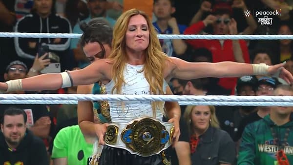 Lyra Valkyria places the championship belt on Becky Lynch, who stands with her arms outstretched in celebration after winning the title at WWE Money in the Bank. Fans are visible in the background, capturing the moment.