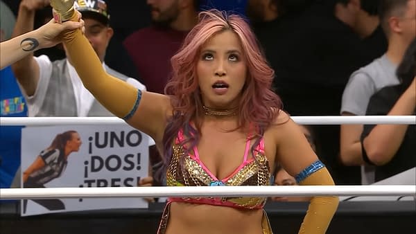 Mina Shirakawa, wearing a sparkling pink wrestling outfit with golden accents, raises her arm in victory after a win on AEW Collision, surrounded by cheering fans.
