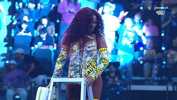 Naomi celebrates her victory in the 2025 Money in the Bank ladder match, wearing a vibrant outfit adorned with money prints and 'Caution' labels, while holding a ladder.