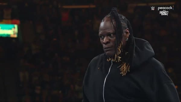 R-Truth, a wrestler with long braided hair, wearing a black hooded sweatshirt, stands in the center of a wrestling ring, with a tense expression on his face, as the crowd is visibly engaged in the background.