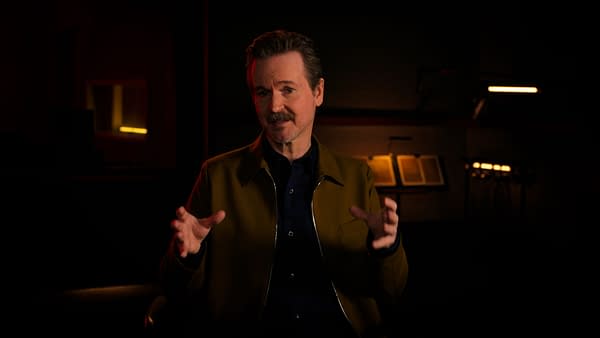 A man with a mustache speaks animatedly, wearing a brown jacket over a dark shirt, in a dimly lit setting with warm lighting. The background features a blurred view of books and instruments.
