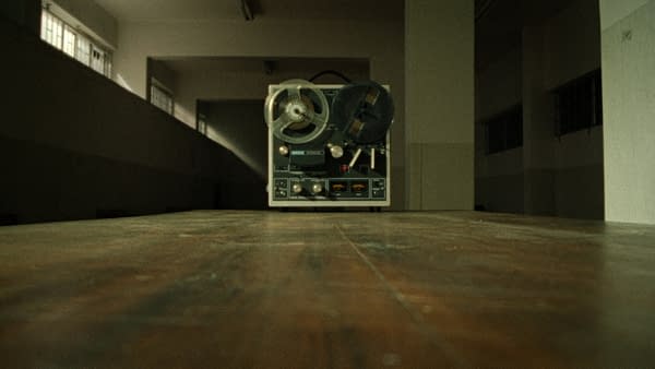 A vintage reel-to-reel tape recorder sits on a wooden table in an empty room with dim lighting. It symbolizes the chilling recordings related to the Son of Sam case.