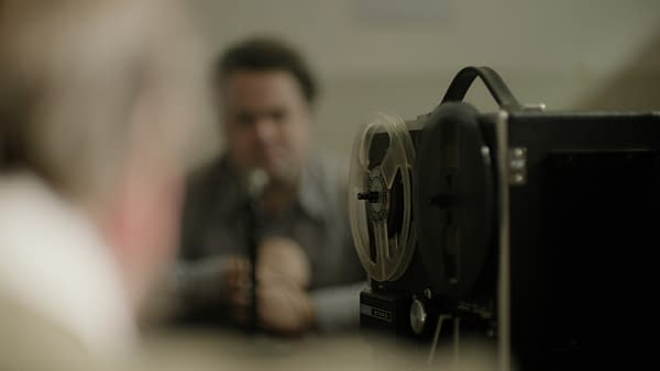 A reel-to-reel tape recorder prominently displayed in the foreground, with a blurred person in the background speaking into a microphone. The setting suggests a historical audio recording session related to criminal investigations.
