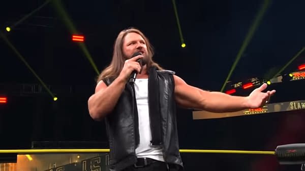AJ Styles stands on stage at TNA Slammiversary, speaking into a microphone and gesturing enthusiastically toward the audience, wearing a black leather vest and tank top.